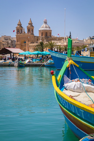 Traditional colourful Maltese Luzzu fishing boats in the turquoise blue water of Marsaxlokk harbour, with the beautiful Parish Church of Our Lady of Pompei, Marsaxlokk, Malta, June 2017のeditorial素材