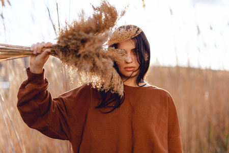 beautiful girl with long dark hair sitting in a beautiful field of flowers on a sunny summer day, holding and looking at some grass flowers in her hand with bright eyesの写真素材