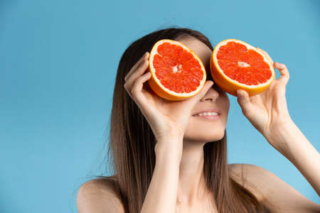 Joyful happy young woman enjoys smell of fresh grapefruit, holding two slices of citrus fruit, cute cheerful girl smiling, posing isolated over blue wall background .の写真素材