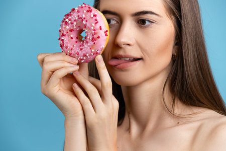 Close-up of a woman with long hair holding a donut and sticking out tongue. Female model a donut in...