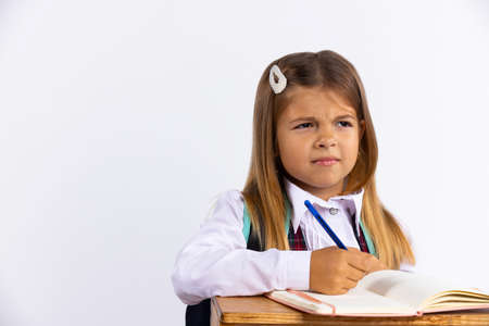 Stressed little girl in school uniform sitting at desk isolated, white background. Schoolgirl unhappy doing homework.の写真素材