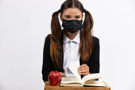 Studio shooting of young woman with black protective mask sitting the desk, flipping through the...