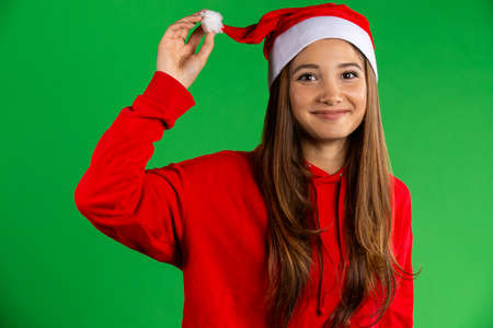 Brunette woman in santa hat and red pullover smiling, looking to the camera on isolated over green backgroundの写真素材