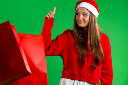 Cute caucasian woman with christmas sale packages, looking far away, pointing up index finger, isolated green backgroundの写真素材