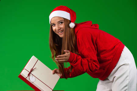 Caucasian smiling brunette woman in santa clothes looks to camera carrying a christmas gift, isolated green backgroundの写真素材