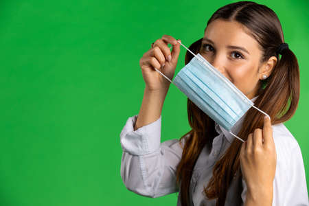 Positive schoolgirl with medical mask. Smiling teen female with ponytails demonstrating disposable medical mask for coronavirus prevention while standing against green backgroundの写真素材