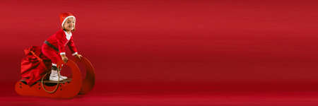 Winter fun BANNER, LONG FORMAT against red background. Babygirl in Xmas clothes and Santas sack drive in imaginary Santa Claus sleigh. Christmas miracle conceptの写真素材