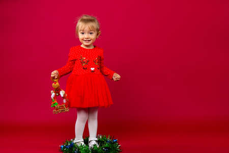 Blonde little girl in red Xmas dress with an reindeer decoration sitting in the middle of the christmas wreath against red background. Merry Christmas . Holiday conceptの写真素材