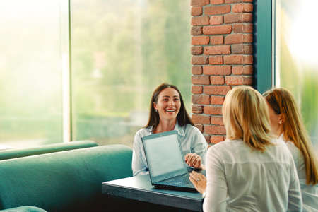 Successful team. Three confident business woman wears white shirts looking at each other and smiling while sitting at the desk in office whith a laptop on the desk .の写真素材