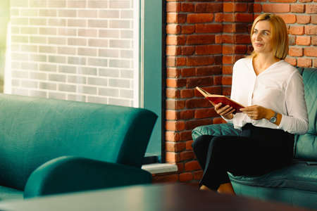 Young confident woman entrepreneur enjoys the work break while sitting on leather sofa in office interior, intelligent awesome female lawyer in formal wear reading a book during work breakの写真素材