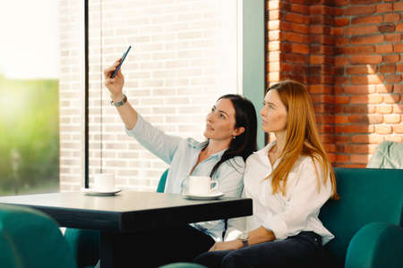 Excited friends make selfie on smartphone having fun in coffeeshop. Students smile for picture on phone met together in cafe. Two young business women posing for self-portrait in office coffee break.の写真素材