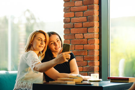 Two women sitting in a restaurant looking at mobile phone and smiling. Friends sitting at a cafe...