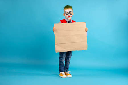 Sad preschool boy at Halloween with Joker face art. The child holds empty protest cardboard with free side space isolated on blue. Social problems and people concept. Preparing for Halloween.の写真素材