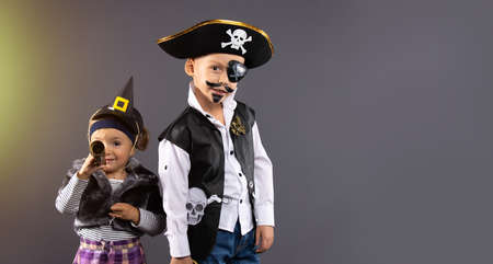 Happy friends, pirat boy and witch little girl at Halloween. Funny kids in carnival costumes on background of gray wall with free side space.Cheerful children ready to celebrateの写真素材