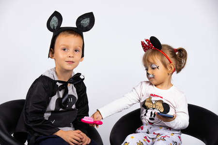 Children in carnival costumes are waiting for the holiday. Boy and girl sitting in black chairs play telephone games, indoors on a white background.の写真素材