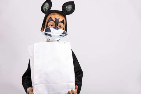Funny halloween concept, little cute boy in bat costume holding a white candy bag with empty space.. Trick or treat. Child in a medical mask on a gray background. The virus attacked the worldの写真素材