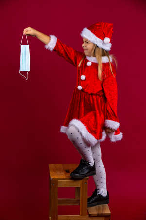 Pretty little santa girl with disgusting face climbs a ladder and wants to decorate something with a protective mask, isolated on red background. New normal concept on winter holidayの写真素材