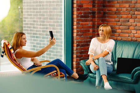 Happy and relaxed female friends sitting on the sofa and rocking chair, in the living room. One takes a selfie while rocking in a chair and the other woman is watching cute. Big city lifeの写真素材