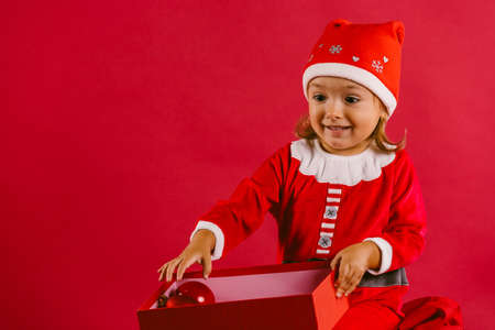 Adorable little girl in a christmas costume with surprised opens a new year gift on a red background with blank spaceの写真素材