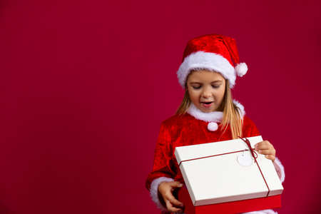Adorable little girl in a Christmas dress wearing a Santa hat opens a gift in surprise on a red background with side empty space. Holidays and winter sale conceptの写真素材