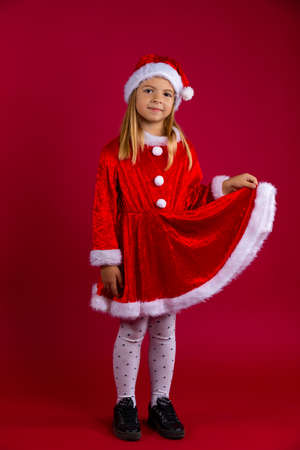 Little girl dressed as Santa Claus and winter hat. Portrait of a child holding a red dress with his hand. Isolated on red backgroundの写真素材