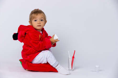 The girl holds her dental kit and looks at the camera, in her pajamas sitting on a pillow. Childhood, faimily, healthy dental care concept on white background with free space.の写真素材
