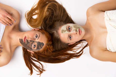 Two girls, blondine and redhaired, girlfriends with different masks on half face, lying on the floor wrapped in towels. Healthy skin and beauty concept on white background. High quality photoの写真素材