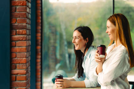 Nice time to relax. Two girls, brunette and redhaired, drink coffee with a smile in sunny weather outside the window. Business office coffee break. High quality photoの写真素材