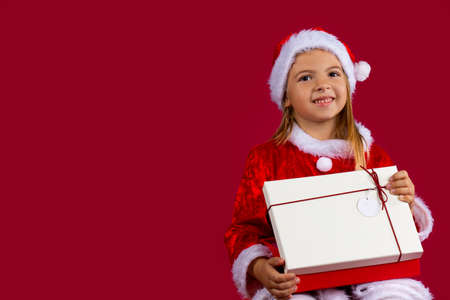 Portrait of a blondine little girl in Santa Claus dress. The girl looks at the camera smiling and opens a gift. On a red isolated background with epty side space. Festive banner. High quality photoの写真素材