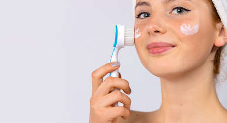 Beauty treatments and facial skin care. A woman with a towel on her head with cream on the cheeks makes herself a massage with cosmetic brush. Photo on white background. High quality photoの写真素材