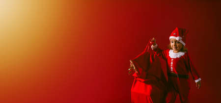 Banner, long format. Little girl in a carnival costume with a sack of Santa Claus on orange to red background, magic corner light. Merry Christmas and a Happy New Year. Holiday Concept. High quality photoの写真素材