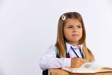 Little girl in a school uniform sitting at a table on a white background with blank space. The schoolgirl is doing her homework and looking away thoughtfully. Education concept. High quality photoの写真素材