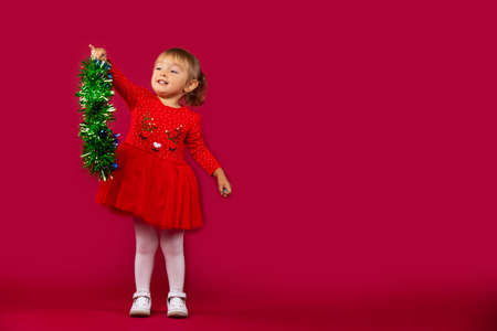 Little happy girl in a red New Year's dress, with a green fluffy garland in her hands. Holiday concept on red background. High quality photoの写真素材