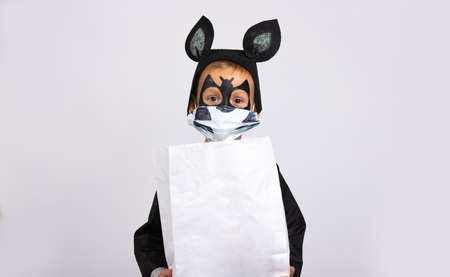 Halloween, little cute boy dressed as a bat holding a white bag of chocolates with empty space .Trick or treat. Child in a medical mask on a gray. The virus has attacked the world. High quality photoの写真素材