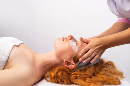 A redhaired girl with patches wrapped in a towel makes a face massage in a cosmetic clinic. Healthy skin concept. Photo on white background. High quality photoの写真素材