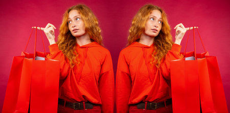 Young redhead girl with freckles and pensive look holding red shopping bags on red background in...