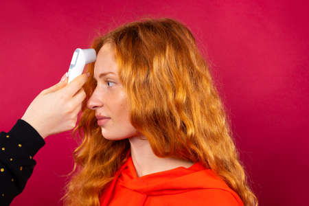 A red-haired girl is measured with non-contact thermometer at her forehead. Photo on a red background. Virus and pandemic concept. High quality photoの写真素材