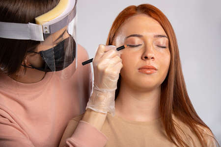 Delicate makeup for a girl with long red hair. Make-up by the master, wearing a mask, gloves and a protective screen to protect against of the virus. Beauty and covid 19 concept. . High quality photoの写真素材