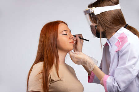 Makeup for a girl with long red hair. New normal beauty concept. Make-up is done by a master in a medical robe, mask, gloves and a protective screen to protect spread of the virus. High quality photoの写真素材