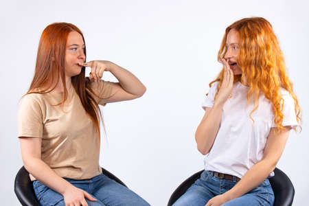 Two red-haired sisters sitting on leather chairs on a white background. Young girls fool around in front of the camera. They make faces and gesture: silence and wow. High quality photoの写真素材