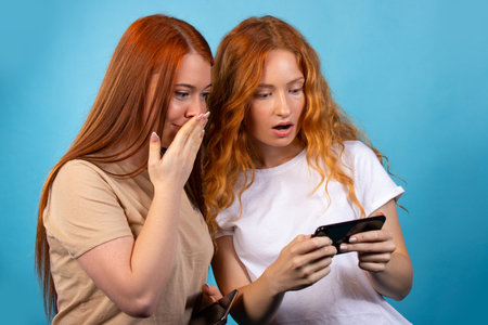 Two red-haired girlfriends in casual clothes. Isolated on blue background. Surprised looking at something on the phone. People lifestyle concept. High quality photoの写真素材