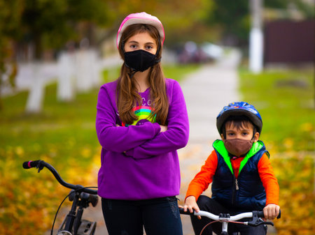 Brother and sister in the autumn park, riding bicycles wearing protective masks. In the background,...