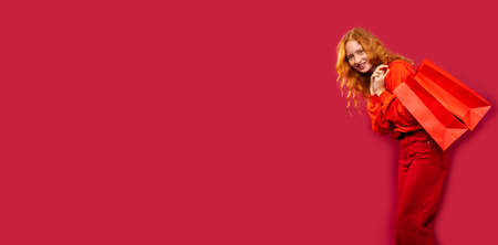 Banner, long format. Happy girl holding red shopping bags on her back on a red background with a lot of empty space. Holiday and shopping concept. High quality photoの写真素材