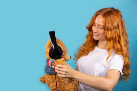 Girl with her beloved bear. The girl put on headphones to the bear, posing and smiling. Photo on a blue background with an empty side spaceの写真素材