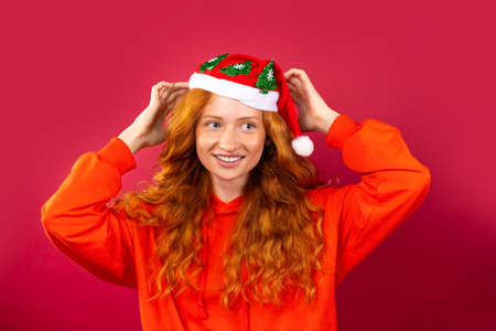 Happy red-haired girl, with a bright smile and gorgeous curly hair, in a Santa Claus hat on a red background. Holiday conceptの写真素材