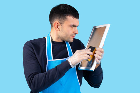 A young man wipes a table mirror to a shine. Gender stereotypes concept. Photo on a blue background....