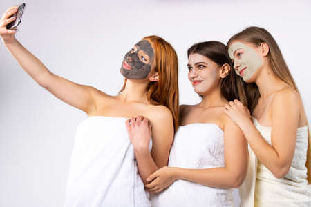 Three young happy girls with facial, wrapped in towels, take a selfie. Beauty and relaxation concept. Photo on white backgroundの写真素材