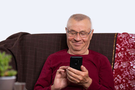 Happy elderly man in glasses holding smartphone while relaxing on the sofa at home. Modern technology concept.の写真素材