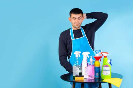 Gender stereotypes. The man holds his head and looks at the set of cleaning products. Photo on a blue background with empty side space.の写真素材