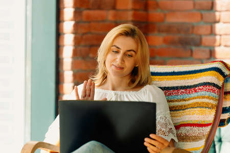 A cute blonde woman communicating by videoconference and waving her hand. Sitting in a comfortable rocking chair.の写真素材